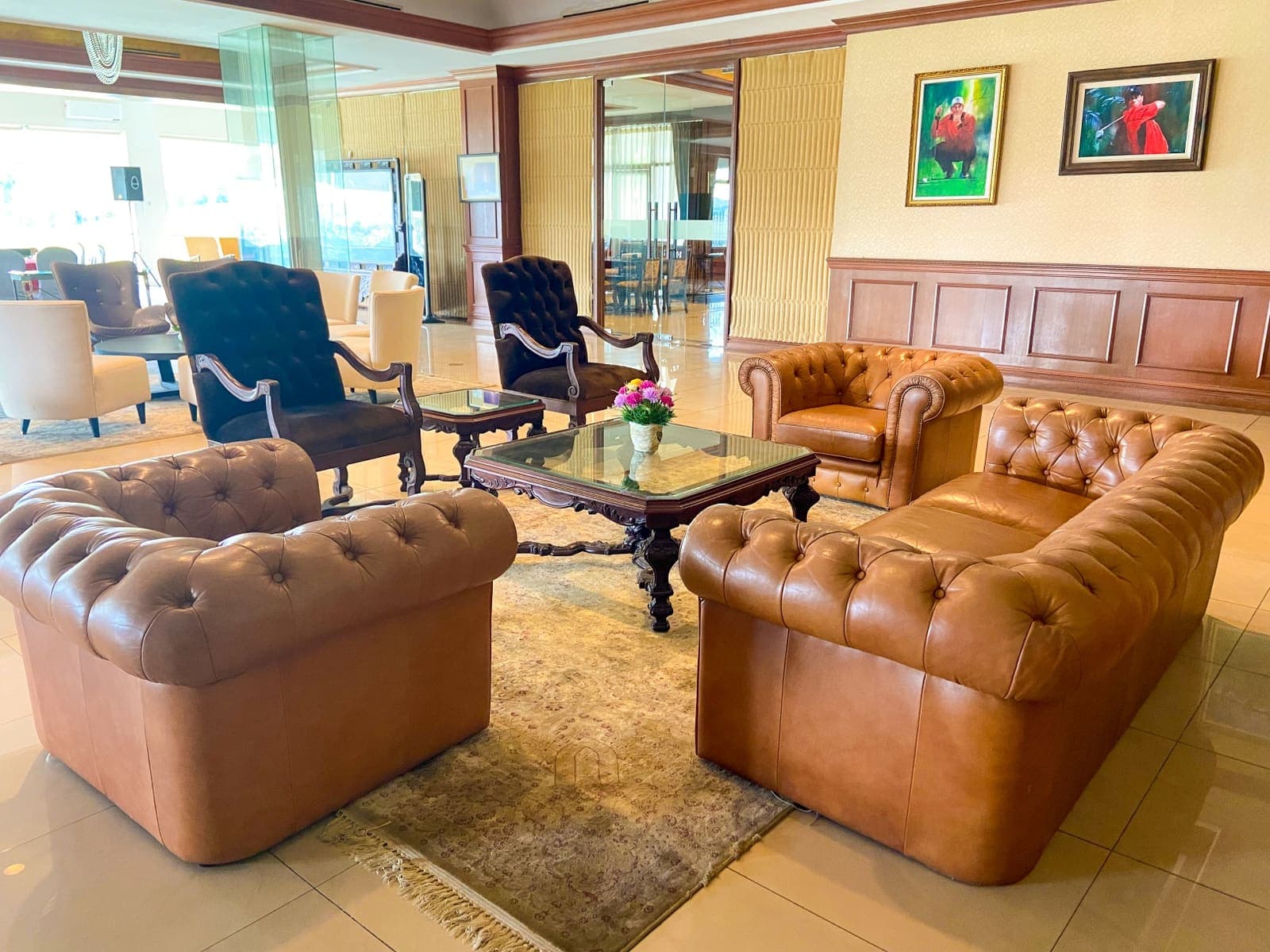 A leather chesterfield lounge with framed golf artwork on the wall and a glass-walled meeting room beyond